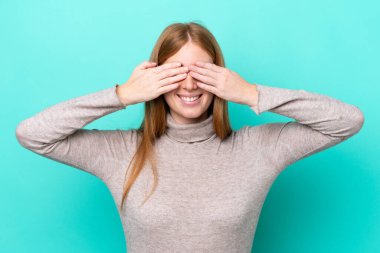 Genç kızıl saçlı kadın mavi arka planda izole edilmiş gözleri elleriyle kaplayan ve gülümseyen.