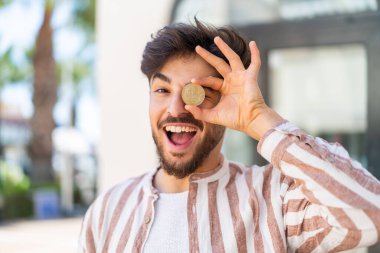 Yakışıklı bir Arap açık havada elinde mutlu bir ifadeyle Bitcoin tutuyor.