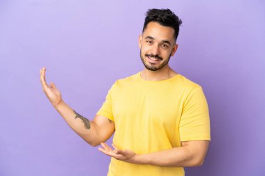 Mor arka planda izole edilmiş genç beyaz adam, davet edilmek için ellerini yana uzatıyor.
