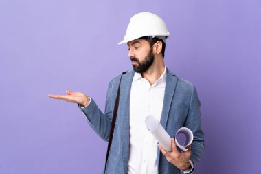 Miğferli genç bir mimar, izole edilmiş mor arkaplan üzerinde planlar tutuyor. Kopyalama alanını şüpheyle tutuyor.