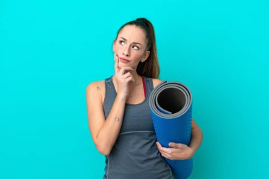 Young sport caucasian woman going to yoga classes while holding a mat isolated on blue background having doubts while looking up