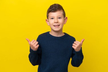 Küçük beyaz çocuk sarı arka planda izole edilmiş. Baş parmakları havada ve gülümsüyor.