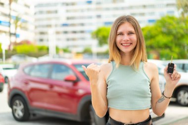 Dışarıda arabanın anahtarlarını tutan genç sarışın kadın bir ürünü sunmak için yan tarafı işaret ediyor.