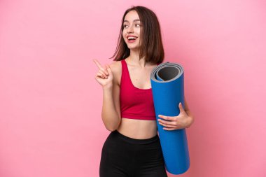 Genç sporcu Ukraynalı kadın yoga derslerine gidiyor pembe arka planda izole edilmiş bir minderi tutarken çözümün farkına varmak için parmağını kaldırıyor.