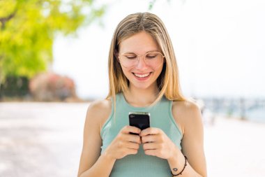 Dışarıdaki genç sarışın kadın cep telefonuyla mesaj ya da e-posta yolluyor.