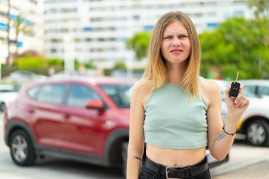 Genç sarışın kadın dışarıda arabanın anahtarlarını tutuyor, üzgün bir ifadeyle.