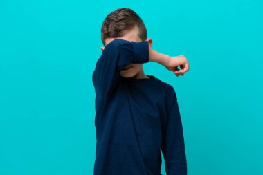Küçük çocuk mavi arka planda izole edildi. Gözlerini elleriyle kapattı.