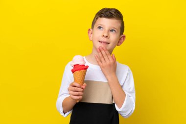 Sarı arka planda izole edilmiş kornet dondurması olan beyaz çocuk gülümserken yukarı bakıyor.