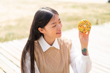 Genç Çinli bir kadın açık havada elinde mutlu bir ifadeyle donut tutuyor.