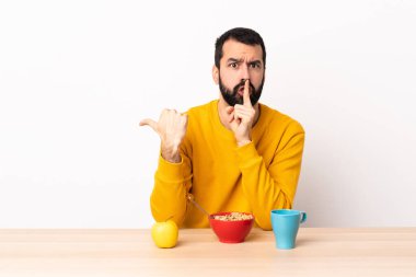 Kafkasyalı bir adam, bir masada kahvaltı yapıyor. Yan tarafı işaret ediyor ve sessiz jest yapıyor..