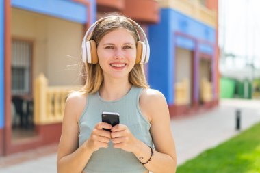 Genç sarışın kadın açık havada müzik dinliyor cep telefonu ve ön tarafa bakıyor.