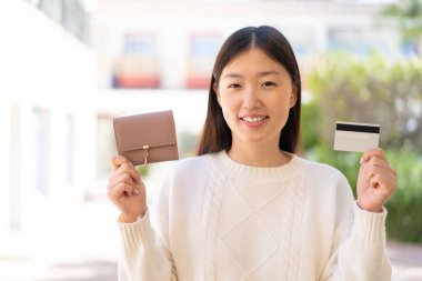 Dışarıdaki güzel Çinli kadın elinde cüzdan ve mutlu bir ifadeyle kredi kartı tutuyor.