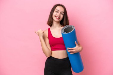 Genç sporcu Ukraynalı kadın yoga derslerine gidiyor. Elinde pembe arka planda izole edilmiş bir minder var. Ürünü sunmak için yan tarafa bakıyor.