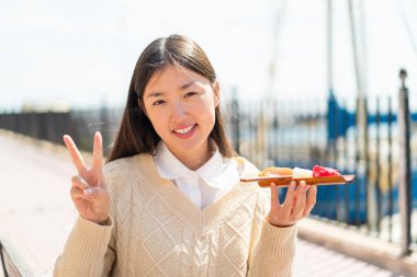Genç Çinli kadın açık havada sashimi tutuyor gülümsüyor ve zafer işareti gösteriyor.
