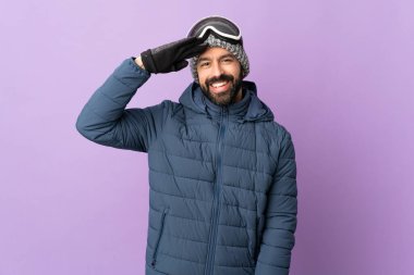 Snowboard gözlüklü kayakçı, izole edilmiş mor arka planda mutlu bir ifadeyle selamlıyor.
