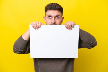 Young caucasian man isolated on yellow background holding an empty placard and hiding behind it