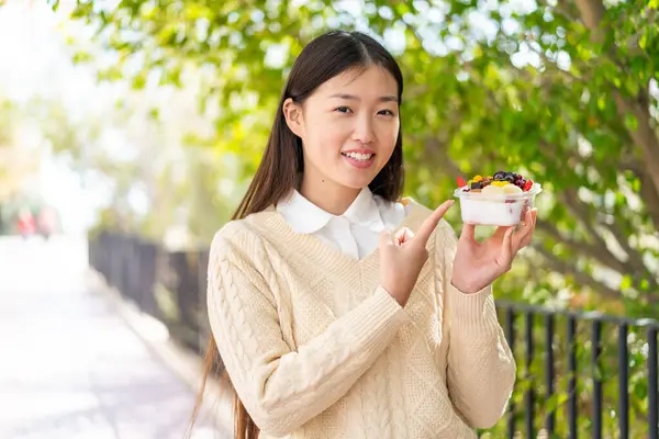 Genç Çinli bir kadın açık havada bir kase meyve tutuyor ve ona doğrultuyor.