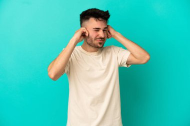 Mavi arka planda izole edilmiş genç beyaz adam hayal kırıklığına uğramış ve kulaklarını kapatmış.