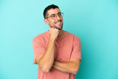 Mavi arka planda soyutlanmış genç, yakışıklı beyaz bir adam. Gözlüklü ve düşünürken yukarı bakıyor.