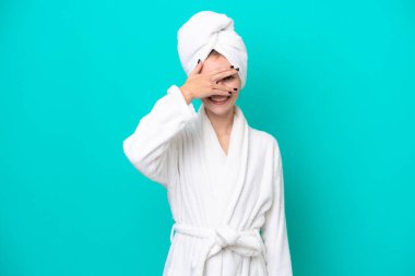 Bornozlu genç bir kadın mavi arka planda izole edilmiş, gözleri kapalı ve gülümsüyor.