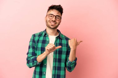 Pembe arka planda izole edilmiş genç beyaz adam bir ürünü sunmak için kenarı işaret ediyor.