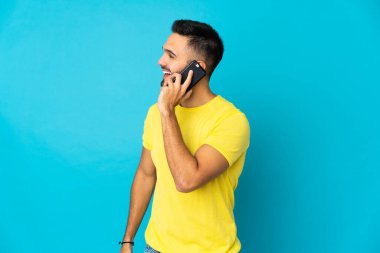 Kafkasyalı genç adam mavi arka planda izole edildi. Cep telefonuyla biriyle sohbet ediyor.