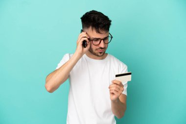 Kafkasyalı genç adam mavi arka planda izole edilmiş cep telefonuyla kredi kartıyla alışveriş yapıyor.