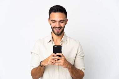 Beyaz arka planda izole edilmiş genç beyaz bir adam cep telefonuyla mesaj ya da e-posta gönderiyor.