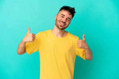 Kafkasyalı genç adam mavi arka planda izole edilmiş bir şekilde baş parmak işareti yapıyor.