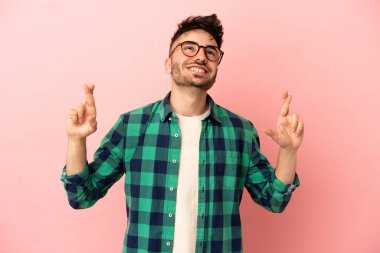 Pembe arka planda izole edilmiş genç beyaz adam parmaklarını çapraz yapıp en iyisini diliyor.