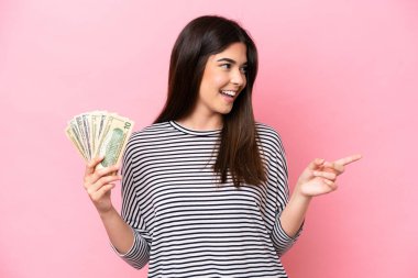 Genç Brezilyalı bir kadın pembe arka planda izole edilmiş bir sürü para alıyor. Parmakla işaret ediyor ve bir ürün sunuyor.