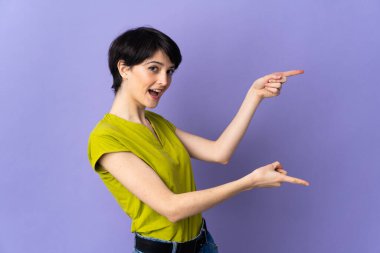 Kısa saçlı bir kadın, mor arka planda izole edilmiş. Parmakla işaret ediyor ve bir ürün sunuyor.