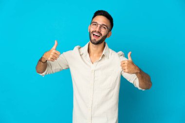 Kafkasyalı genç adam mavi arka planda izole edilmiş bir şekilde baş parmak işareti yapıyor.