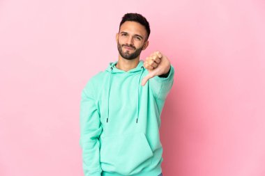 Pembe arka planda izole edilmiş genç beyaz adam olumsuz ifadeyle baş parmağını aşağı gösteriyor.