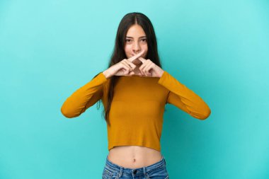 Mavi arka planda izole edilmiş genç bir Fransız kadın sessizlik işareti gösteriyor.