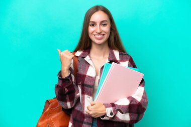 Mavi arka planda izole edilmiş genç beyaz kadın bir ürünü sunmak için kenarı işaret ediyor.