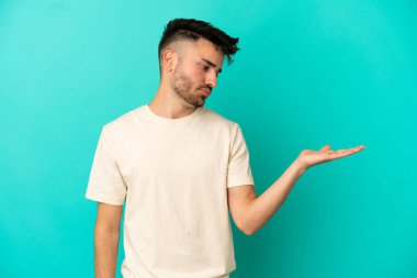 Mavi arka planda izole edilmiş genç beyaz adam, kopyalama alanını şüphe ile tutuyor.