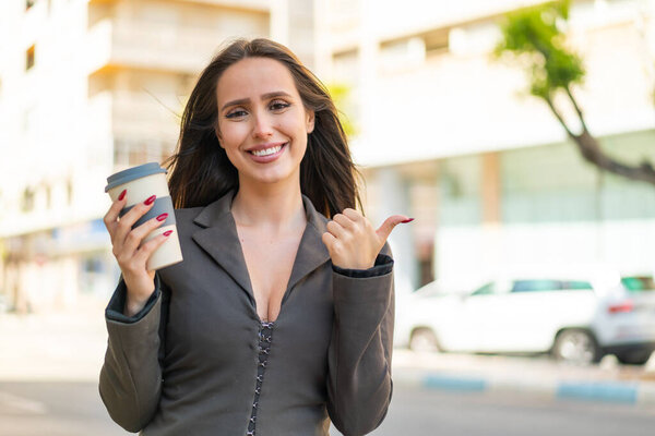 Young woman holding a take away coffee at outdoors pointing to the side to present a product