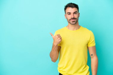 Mavi arka planda izole edilmiş genç beyaz adam bir ürünü sunmak için kenarı işaret ediyor.