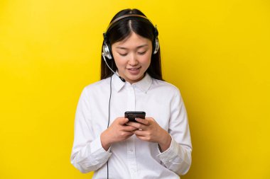 Telefon pazarlamacı Çinli kadın sarı arka planda izole edilmiş bir kulaklıkla cep telefonuyla bir mesaj gönderiyor.