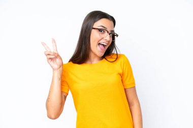 Beyaz arka planda izole edilmiş genç beyaz kadın gülümsüyor ve zafer işareti gösteriyor.