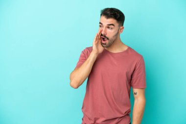 Mavi arka planda izole edilmiş genç beyaz adam yan tarafa bakarken sürpriz bir hareketle bir şeyler fısıldıyor.