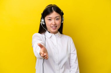 Tele pazarlamacı Çinli bir kadın sarı arka planda izole edilmiş bir kulaklıkla çalışıyor ve iyi bir anlaşma için el sıkışıyor.