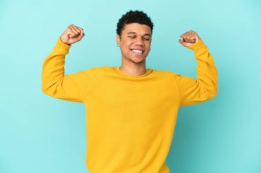 Genç Afrikalı Amerikalı adam mavi arka planda izole edilmiş, güçlü bir jest yapıyor.