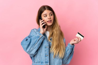 Mavi arka planda izole edilmiş genç sarışın kadın cep telefonuyla sohbet ediyor ve elinde bir kredi kartı tutuyor.