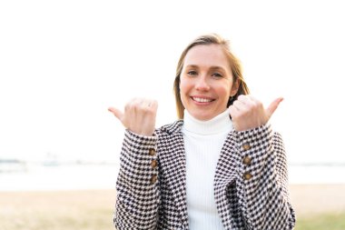 Açık havada genç, sarışın bir kadın. Baş parmakları havada ve gülümsüyor.