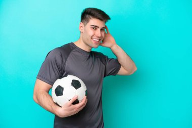 Kafkasyalı genç futbolcu mavi arka planda izole edilmiş bir şekilde elini kulağına koyarak bir şeyler dinliyor.