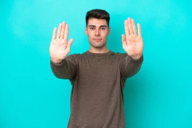 Mavi arka planda izole edilmiş genç beyaz adam dur hareketi yapıyor ve hayal kırıklığına uğruyor.