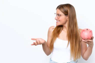 Genç, sarışın bir kadın kumbarayı elinde tutuyor beyaz arka planda, şaşırmış bir yüz ifadesi var.