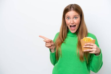 Beyaz arka planda izole edilmiş kızarmış patates kızartması tutan genç beyaz kadın şaşırmış ve parmağıyla kenarı işaret ediyor.